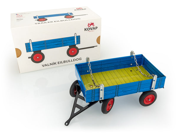 A Kovap toy trailer for a tractor with a folding tailgate, predominantly blue with red wheels, displayed alongside its packaging box.