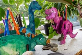Multiple Brachiosaurus-shaped pen pot with multiple compartments, filled with pencils and other stationery items. The pen pots are blue, pink and green.