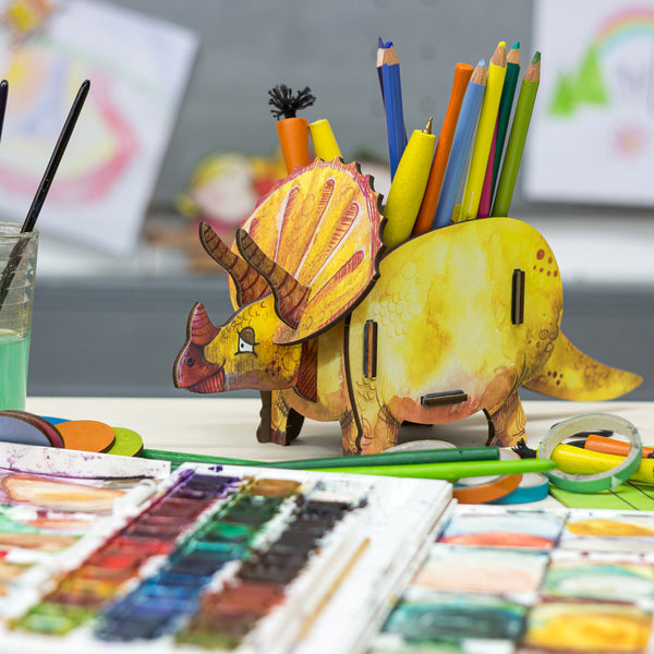 wooden pen pot in the shape of a yellow triceratops dinosaur with colourful paints next to it